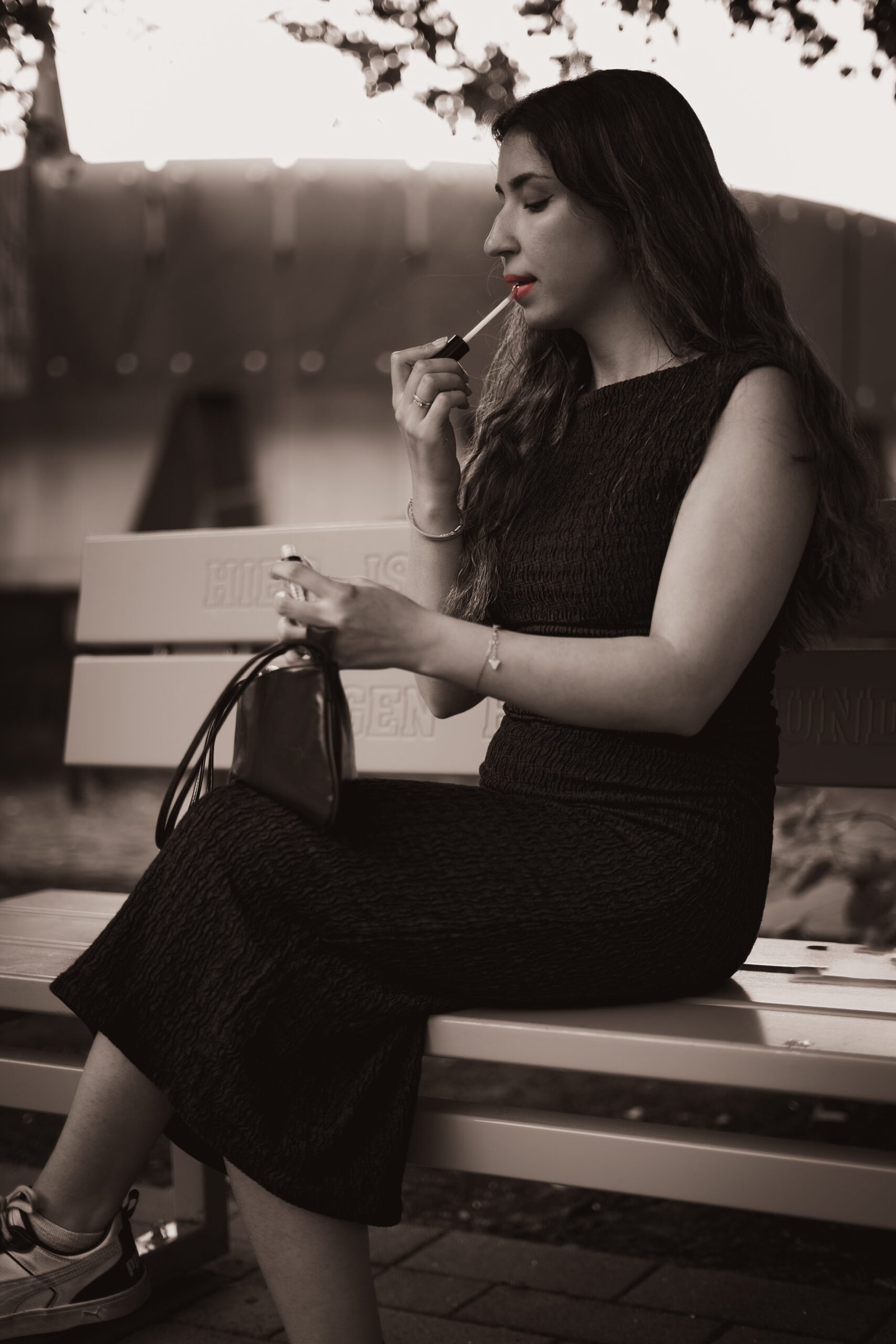 Kreatives Colorkey-Portrait in Schwarz-Weiß: Eine junge Frau in einem schwarzen, strukturierten Kleid sitzt im Freien auf einer Bank. Sie hält eine Handtasche auf dem Schoß und trägt gerade Lippenstift auf. Das gesamte Bild ist monochrom in Sepiatönen gehalten, nur ihre Lippen und der Applikator des Lippenstifts leuchten in einem kräftigen Rot. Professionelle Portraitfotografie aus Osnabrück.
