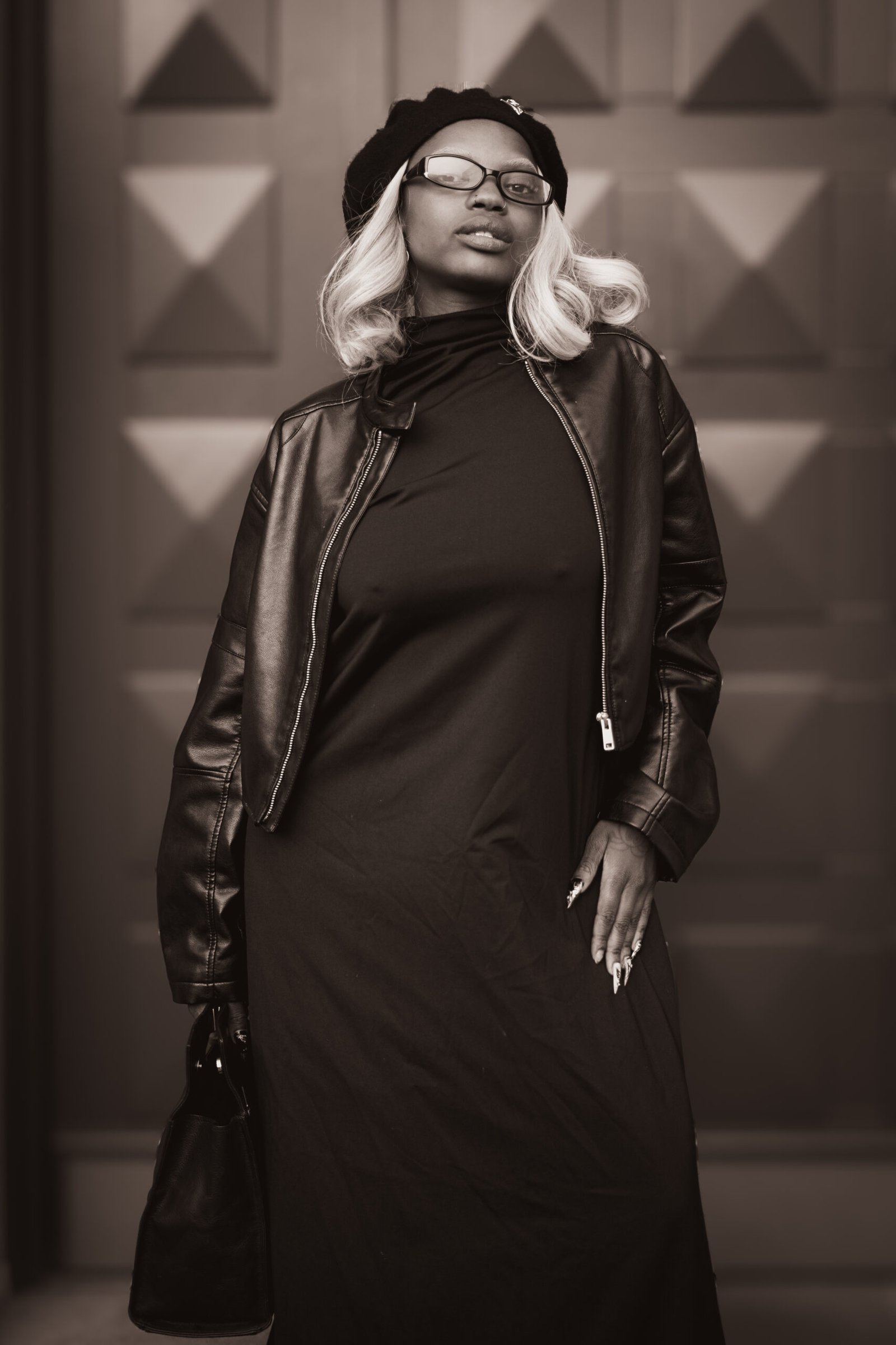 Ein stilvolles Fashion-Portrait in Schwarz-Weiß (Sepia-Look). Eine junge Frau mit platinblonden Haaren posiert selbstbewusst in einem komplett schwarzen Outfit, bestehend aus einem langen Kleid, einer Lederjacke und einer Baskenmütze. Sie trägt eine Brille und hält eine Ledertasche. Im Hintergrund ist ein dunkles Tor mit geometrischem Rautenmuster zu sehen. Professionelles Streetstyle-Shooting aus Osnabrück.