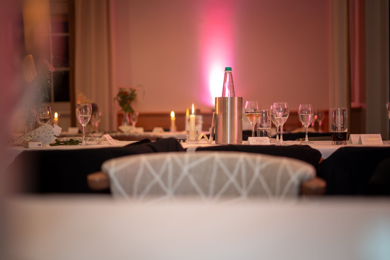 Stimmungsvolle Tischdekoration auf einer Hochzeitsfeier in Osnabrück. Detailaufnahme eines festlich gedeckten Tisches mit Weingläsern, brennenden Kerzen, Namenskärtchen und einer eleganten rosa Hintergrundbeleuchtung im Saal.