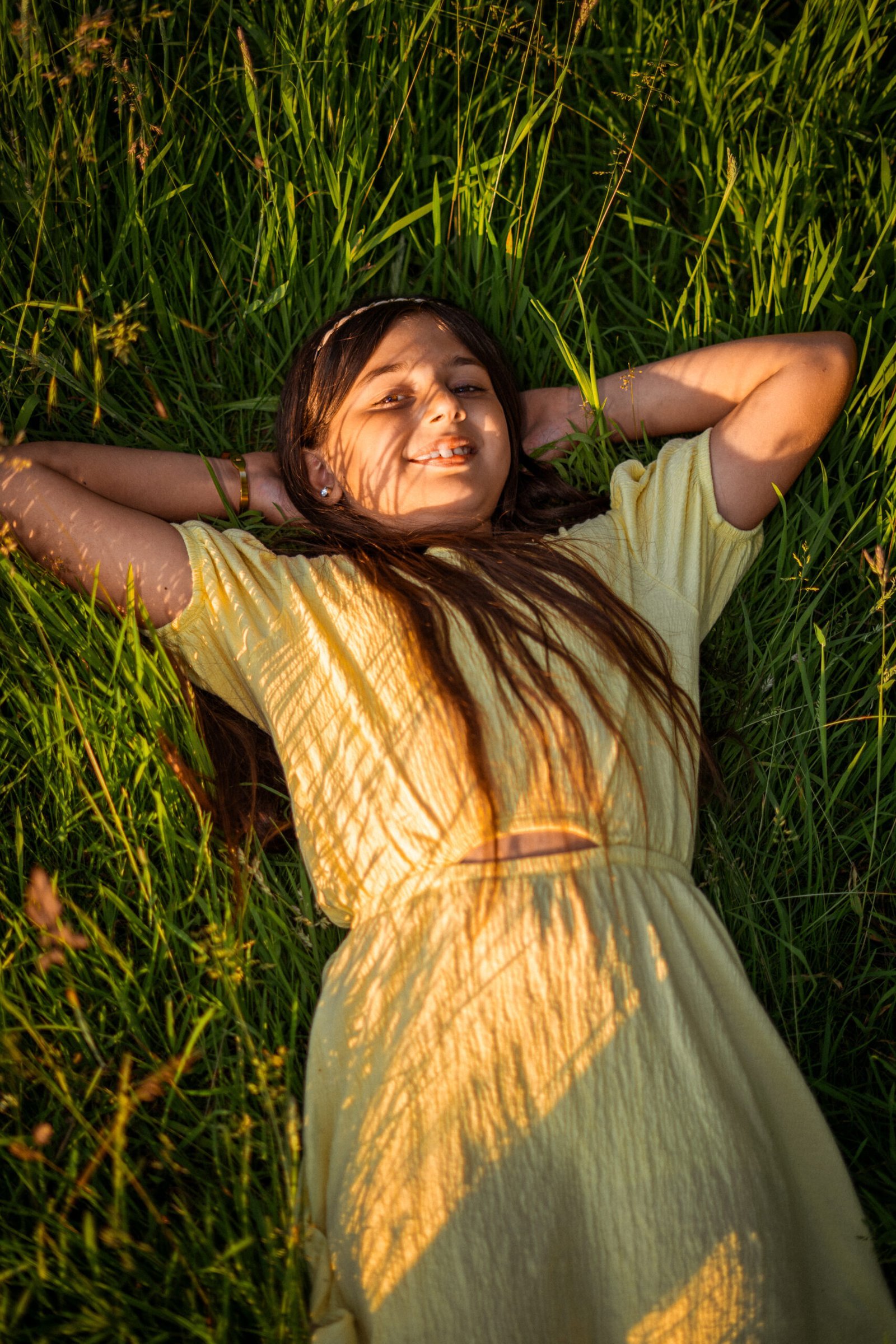 Ein idyllisches Kinderportrait im warmen Licht der Golden Hour. Ein Mädchen in einem hellgelben Sommerkleid liegt entspannt auf dem Rücken in hohem, grünem Gras, die Arme hinter dem Kopf verschränkt. Schöne Schatten von Grashalmen fallen auf ihr Gesicht und Kleid. Professionelle Outdoor-Kinderfotografie in Osnabrück.