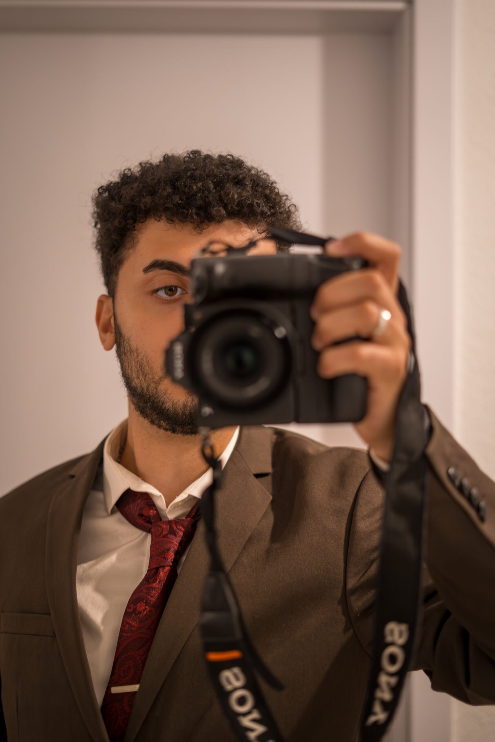 Portrait von Mirovisuals, dem Hochzeits- und Eventfotografen aus Osnabrück. Er trägt einen eleganten braunen Anzug mit roter Krawatte und fotografiert sich selbst im Spiegel mit einer Sony-Kamera.