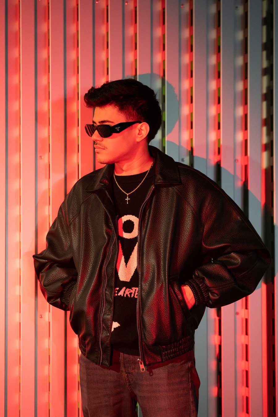 Kreatives Fashion-Portrait eines jungen Mannes im Streetwear-Look. Er trägt eine schwarze Lederjacke, einen Pullover mit Print und eine dunkle Sonnenbrille und schaut cool zur Seite. Er steht vor einer strukturierten Wand, die von intensivem rotem Neonlicht angestrahlt wird, was starke Kontraste erzeugt. Professionelle Studiofotografie aus Osnabrück.