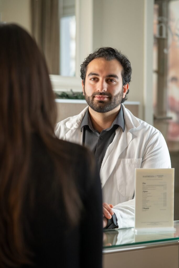 Vertrauensvolles Beratungsgespräch in einer Klinik für ästhetische Medizin. Ein männlicher Arzt im Kittel hört einer Patientin aufmerksam zu. Authentische Corporate-Fotografie für Arztpraxen und Kliniken in Osnabrueck.