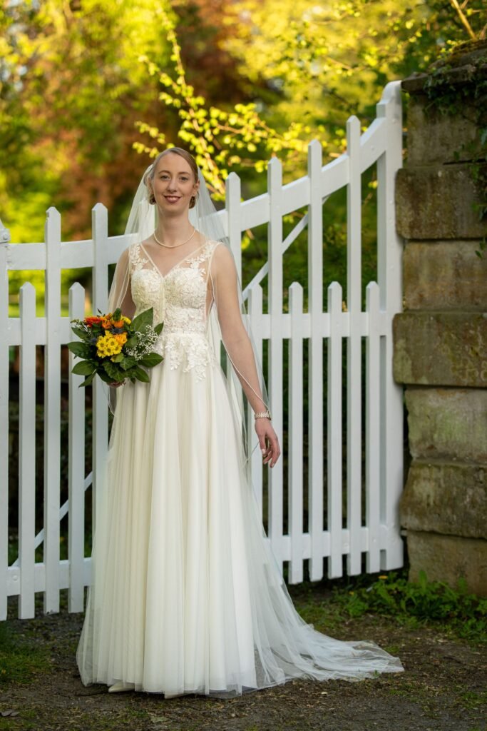 Elegantes Braut-Shooting an der Wasserburg Schelenburg: Braut im Spitzenkleid mit Schleier und buntem Brautstrauß vor einem weißen Holztor in Bissendorf. Professionelle Hochzeitsfotografie aus Osnabrück.
