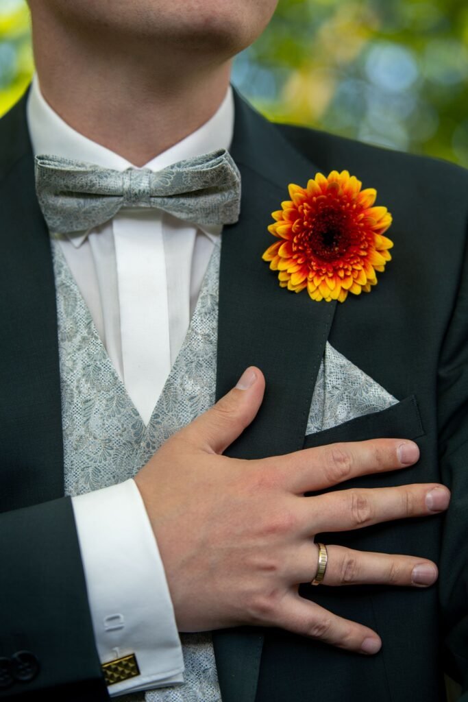 Detailaufnahme der Hochzeitsgarderobe: Bräutigam mit goldener Ansteckblume, Ehering und gemusterter Fliege an der Wasserburg Schelenburg. Elegante Hochzeitsfotografie aus Osnabrück.