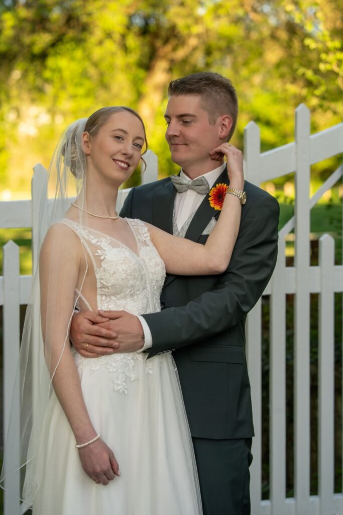Inniges Brautpaar beim Shooting vor einem weißen Gartenzaun an der Wasserburg Schelenburg in Bissendorf. Professionelle Hochzeitsfotografie für Osnabrück und das Osnabrücker Land für authentische Momente.