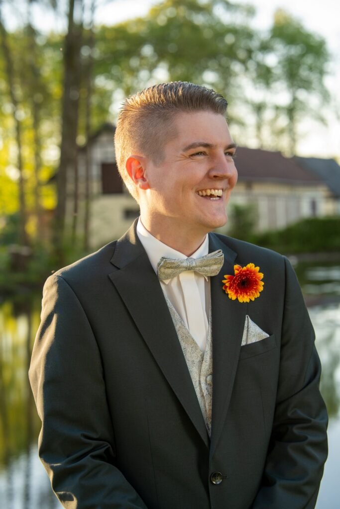 Authentisches Portrait eines fröhlich lachenden Bräutigams im Anzug mit orangefarbener Blume am Revers. Aufgenommen im Park der Schelenburg in Bissendorf bei Osnabrück vom Hochzeitsfotograf Osnabrück