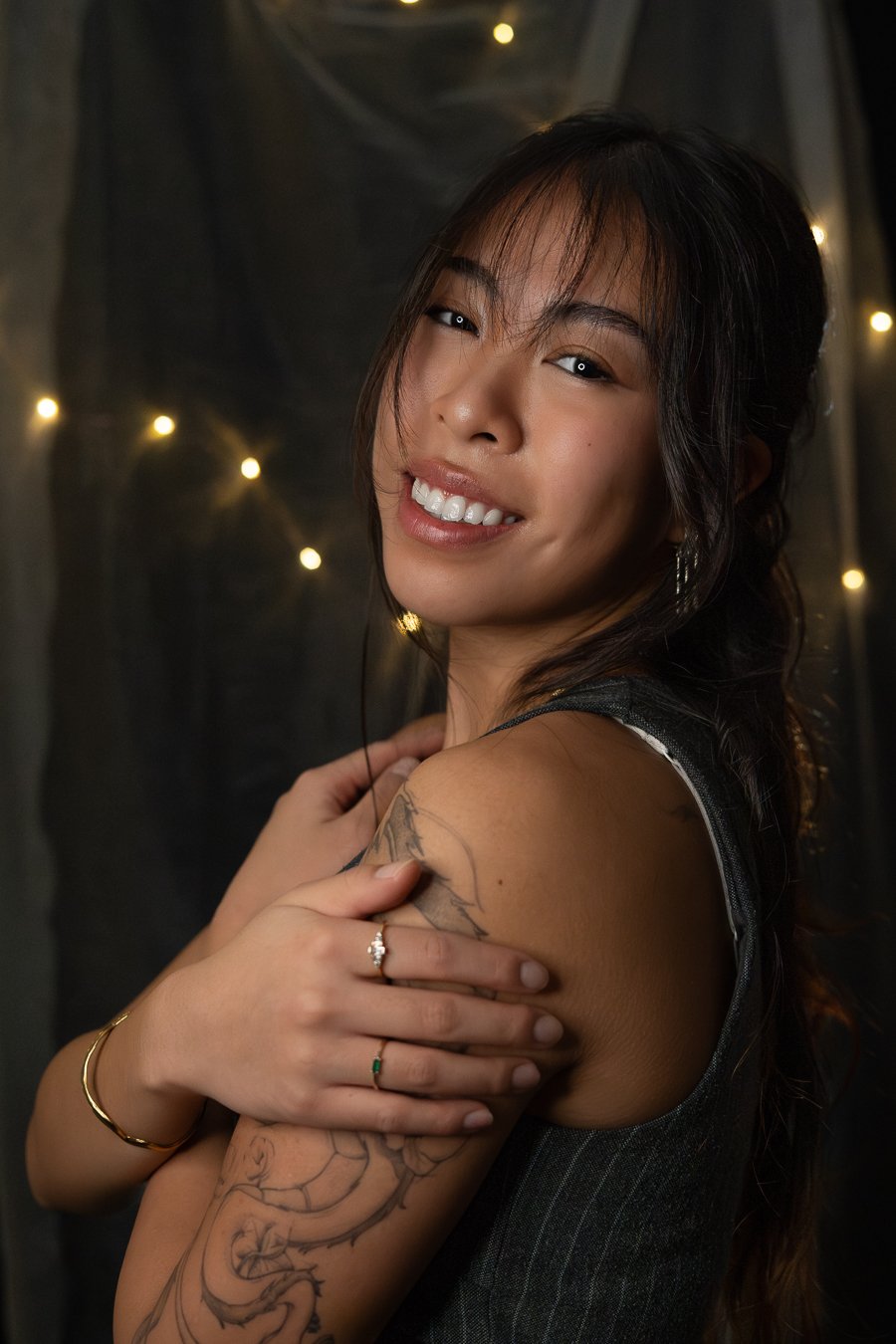Authentisches Lifestyle-Porträt einer lächelnden Frau mit Drachen-Tattoo am Oberarm vor einem stimmungsvollen Hintergrund mit Lichterketten, aufgenommen von Mirovisuals, deinem Fotografen für Porträts in Osnabrück.