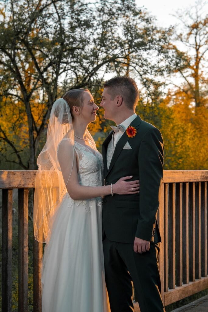 Braut im Spitzenkleid und Bräutigam im grünen Anzug stehen sich lächelnd auf einer Brücke gegenüber. Warmes Abendlicht an der Schelenburg, aufgenommen von einem professionellen Hochzeitsfotografen aus Osnabrück.