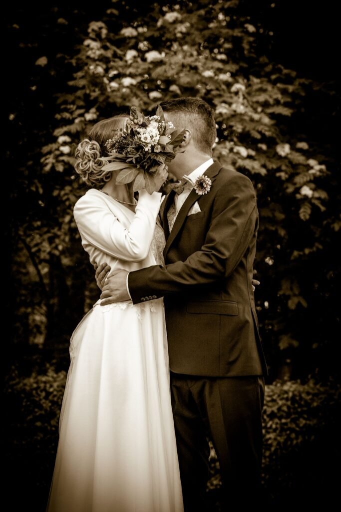 Kreatives Brautpaar-Portrait in Sepia-Optik: Ein Kuss hinter dem Blumenstrauß im Park der Schelenburg. Zeitlose Hochzeitsbilder im Osnabrücker Land vom Hochzeitsfotograf Osnabrück.